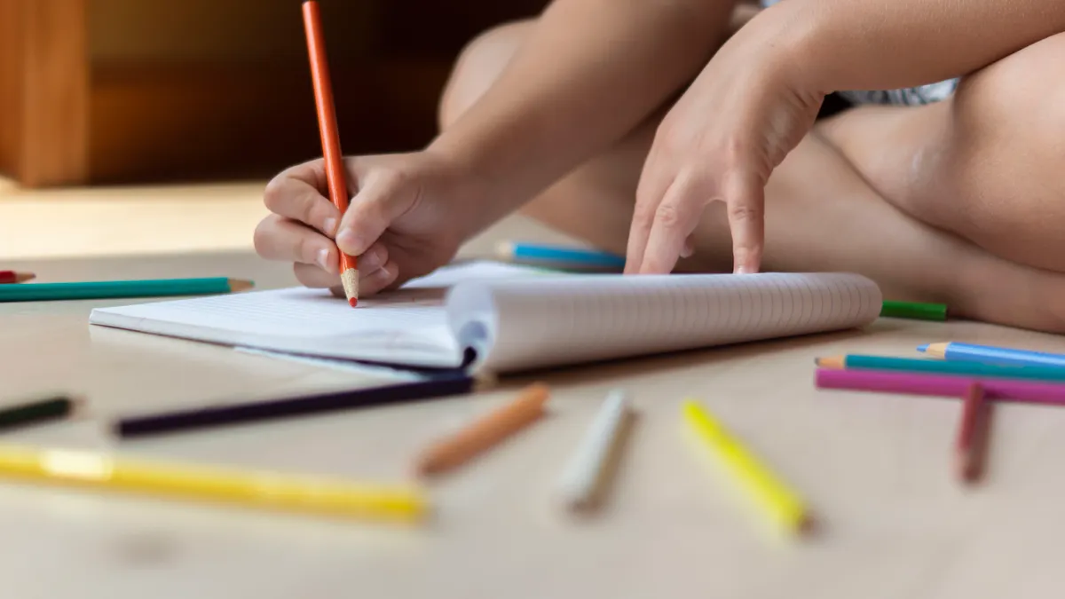 person writing in a notebook with pencils nearby